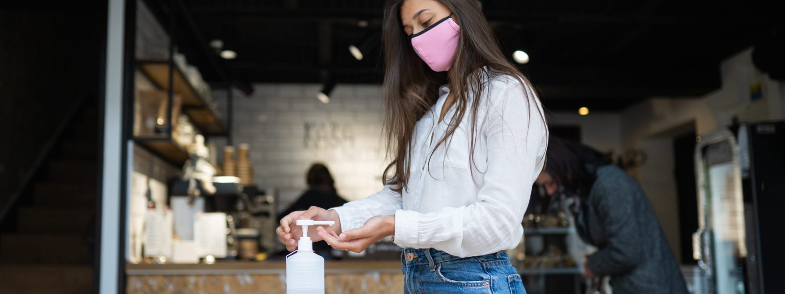 Young beautiful woman with attractive smile in protective mask using hand sanitizer gel to wash her hands. Girl stands threshold outside in cafe scene. Corona Virus prevention concept.
